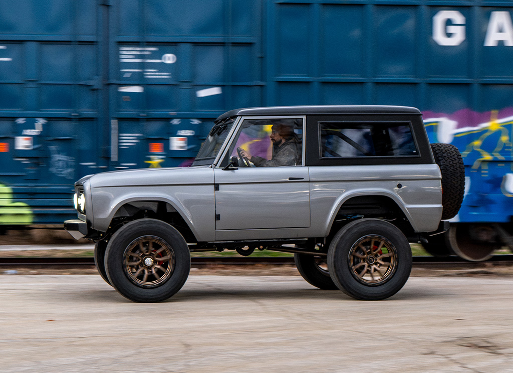 Restored gray 1972 Ford Bronco - side profile view driving by a train car.